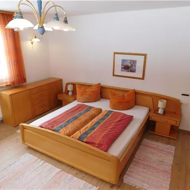 Ein gemütliches Schlafzimmer mit einem großen Bett und bunten Kopfkissen. Der Raum ist hell mit Holz- und Stoffelementen gestaltet.