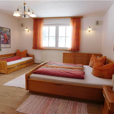 Ein helles Schlafzimmer mit zwei Betten, einem großen Fenster und orangefarbenen Vorhängen. Der Raum enthält Holzmöbel und eine gemütliche Atmosphäre.