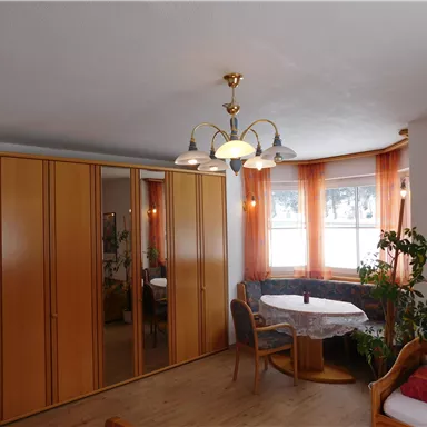Ein helles Zimmer mit einem großen Spiegelkleiderschrank und einem runden Tisch. Die Fenster sind mit orangen Vorhängen geschmückt und es gibt Pflanzen im Raum.