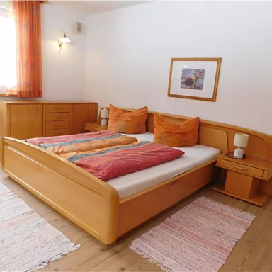 Ein gemütliches Schlafzimmer mit einem Doppelbett und Holzmöbeln. Die Fenster sind mit orangefarbenen Vorhängen dekoriert und es gibt einen Nachttisch mit einer Lampe.