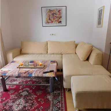 Ein gemütliches Wohnzimmer mit einer hellen Couch und einem Couchtisch. Auf dem Tisch stehen Kerzen und die Wände sind mit Kunstbildern dekoriert.