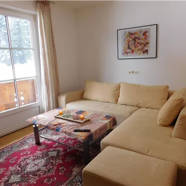 Ein gemütliches Wohnzimmer mit einem beigen Sofa und einem kleinen Tisch. Im Hintergrund sind ein Fenster und eine Balkontür, die einen Blick auf den Schnee bieten.