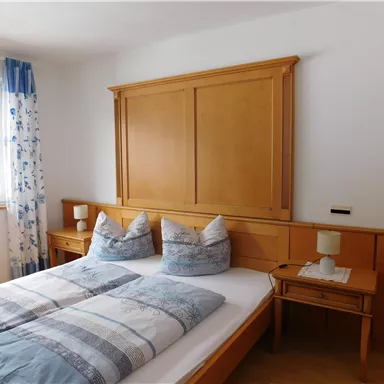 Ein helles Schlafzimmer mit einem Doppelbett und einer Holzverkleidung. Die Fenster sind mit blauen Vorhängen dekoriert und ein Nachttisch steht neben dem Bett.