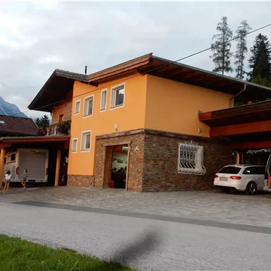 A modern house with an orange facade and stone cladding. In the foreground, cars are parked in the driveway.