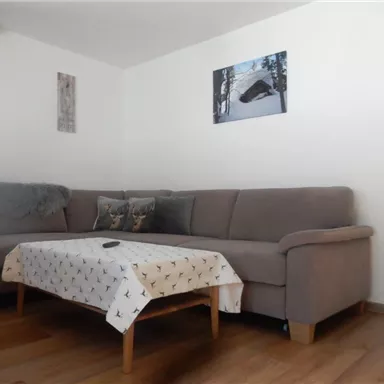 A cozy living room setup with a large sofa and a table. Pictures hang on the wall, and there is a carpet on the floor.