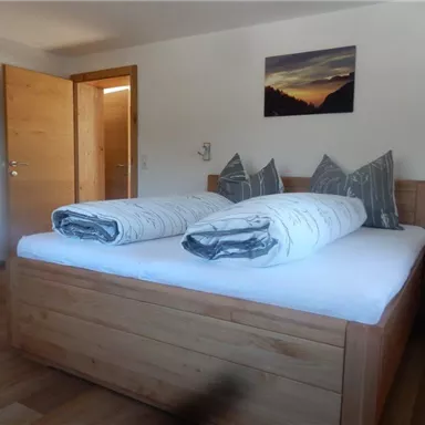 A simple bedroom with a large wooden bed and two pillows. The walls are light and a door leads to another room.