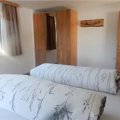 A bright bedroom with a window and a wooden wardrobe. Thick blankets with a floral pattern are on the bed.