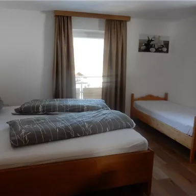 A simple bedroom with two beds and a single bed. The room has large windows and bright curtains.