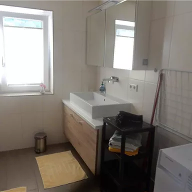 A modern bathroom with a white vanity unit and a large mirror. There is a window, a washing machine, and a laundry rack next to colorful towels.