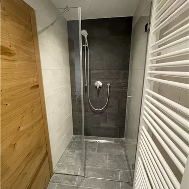 A modern shower with a glass wall and gray tiles. In the background, a towel radiator and a wooden door are visible.