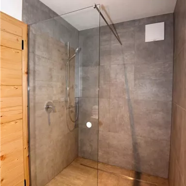 A modern shower cabin with a glass partition and gray tiled walls. The floor is made of light material, and the wooden details add a warm touch.