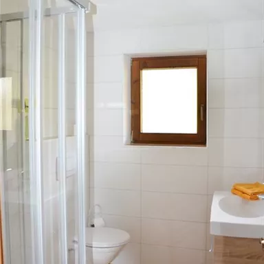 A modern bathroom with a shower and a toilet. The room has light tiles and a window with a wooden frame.
