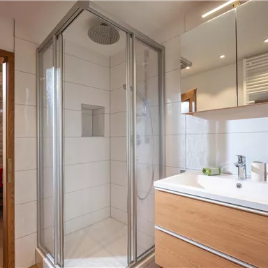 A modern bathroom with a walk-in shower and an elegant wooden sink. The room is bright and friendly, with mirrors and soft lighting.