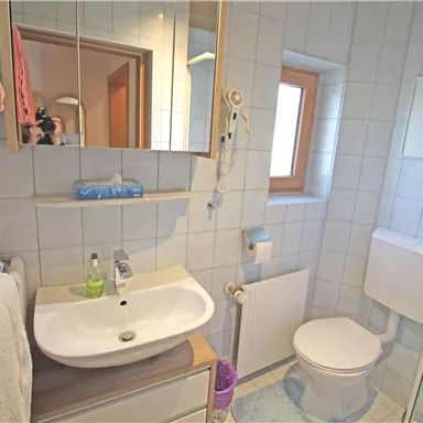 A bright bathroom with a vanity, sink, and shower. The walls are tiled and there is a window that lets in natural light.