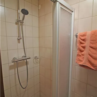 A simple shower in a bathroom with white tiles. An orange towel hangs next to the shower.