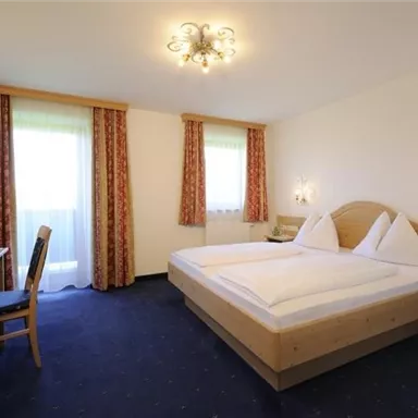 A cozy bedroom with a large double bed and wooden furniture. The room has large windows that let in plenty of daylight.
