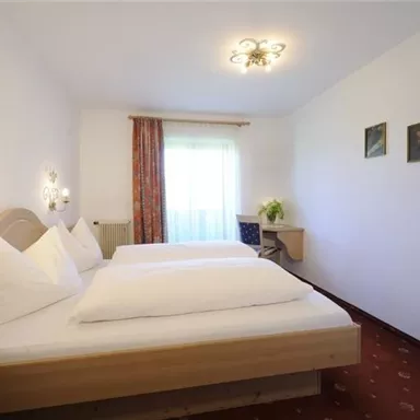 A cozy bedroom with two beds and a bright window front. The walls are in neutral colors and the furnishings are simple and inviting.