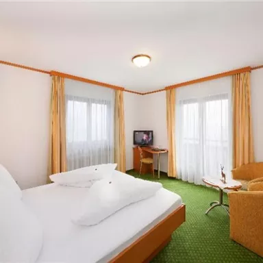 A cozy hotel room with a double bed and a small table. Large windows let in plenty of light, and the green carpeted area creates a pleasant atmosphere.