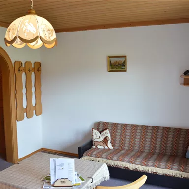 A cozy living room with a sofa, a table, and a television on a shelf. The walls are light, and a beautiful lamp hangs from the ceiling.