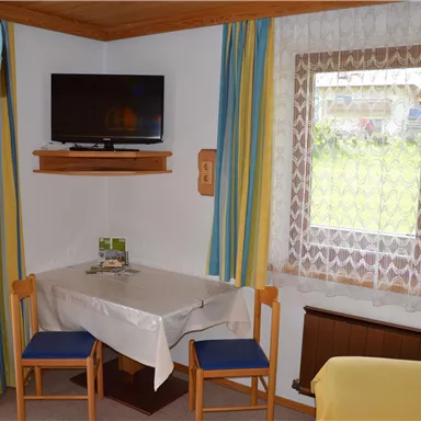 A cozy living space with a table and two chairs. A television hangs on the wall and a window lets in plenty of daylight.
