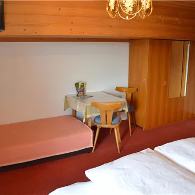 A cozy room with wooden walls, a double bed, and a small dining table. In the background, there is an additional sleeping sofa.