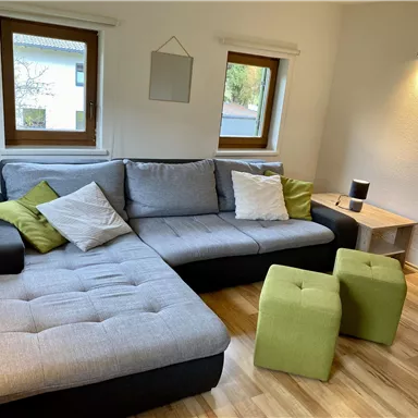 Ein modernes Wohnzimmer mit einer bequemen Couch und Sitzhockern. Die vier Fenster lassen viel Tageslicht herein.