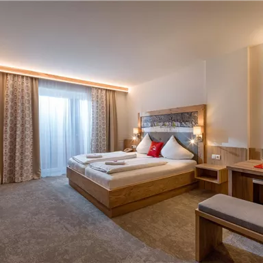 A modern hotel room with a double bed and a cozy seating area.  
Large windows let in plenty of daylight and the decor is made of light wood.