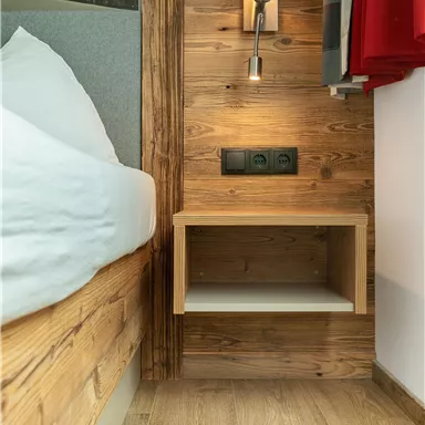 A cozy bedroom with a wooden bed and a bedside lamp.
On the wall, there are power outlets and a small table.