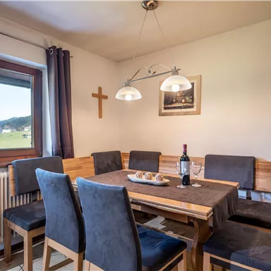 An inviting dining area with a wooden table and six chairs. Large windows offer views of the landscape, and a bottle of wine sits on the table.