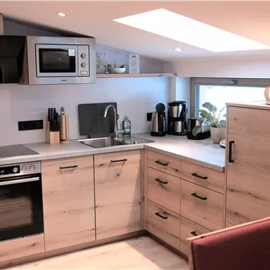 A modern kitchen with wooden surfaces and new appliances. It features a stove, a sink, and a microwave, as well as a view to the outside.