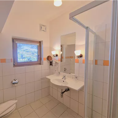 A bright bathroom with a shower, sink, and mirror. It has white tiles and a window that lets in light.