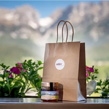 A brown paper bag is next to a glass with a cream. In the background, there are blooming plants and mountains visible.