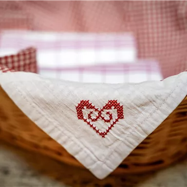 A basket with an embroidered heart motif on the blanket. Inside, there are several checkered patterned cloths.