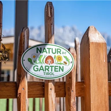 A sign on a wooden fence with the inscription "NATURE GARDEN TIROL". In the background, mountains and a clear blue sky can be seen.