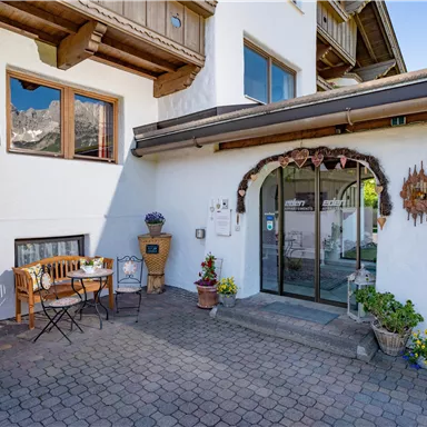 An inviting entrance area of a house with a glass door and colorful flowers. There is a small table with chairs, surrounded by a cozy atmosphere.