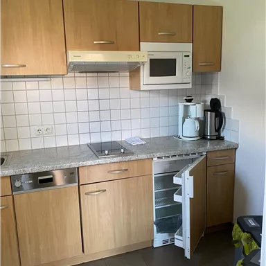 A modern kitchen with wooden cabinets and white tiles. There is a refrigerator, a microwave, and common kitchen appliances.
