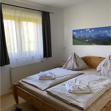 A bright bedroom with two single beds and a large window. The walls are painted in neutral colors, and a landscape painting adorns the wall.