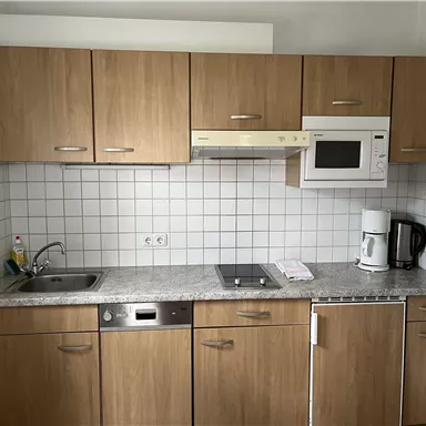 A modern kitchen with wooden cabinets, a sink, and a microwave oven. The countertop is made of gray stone and there are various kitchen appliances.