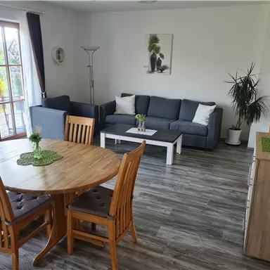 Ein modernes Wohnzimmer mit einer gemütlichen Couch und einem Holztisch. Große Fenster lassen viel Licht herein und es sind Pflanzen dekorativ platziert.