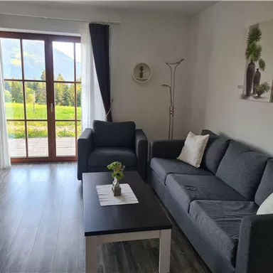 Ein helles Wohnzimmer mit einem grauen Sofa und einem Tisch. Große Fenster bieten einen Blick auf die grüne Landschaft draußen.