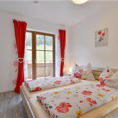 Ein gemütliches Schlafzimmer mit zwei Betten, dekoriert mit Blumenbettwäsche. Große Fenster mit roten Vorhängen bieten einen Blick nach draußen.