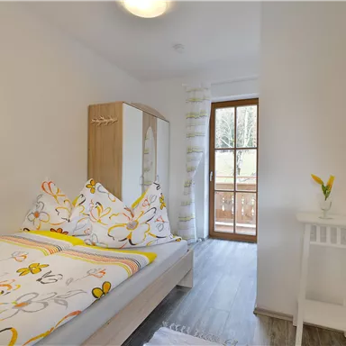 Ein helles Schlafzimmer mit einem Bett und bunten Bettbezügen. Fenster mit Aussicht auf einen Balkon und dekorative Elemente an den Wänden.