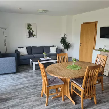 Ein modernes Wohnzimmer mit einer gemütlichen Couch und einem Esstisch aus Holz. Die Wände sind hell und die Einrichtung ist einfach und einladend.
