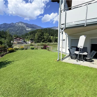 A green meadow with a small garden area and a table with chairs. In the background, there are mountains and a blue sky with a few clouds.