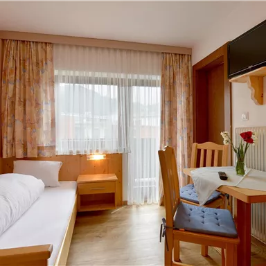 Ein gemütliches Zimmer mit einem Einzelbett, einem Tisch und Stühlen. Große Fenster sorgen für viel Tageslicht.
