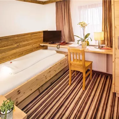 Ein gemütliches Schlafzimmer mit Holzmöbeln und einem Einzelbett. Am Arbeitsplatz steht ein Fernseher, und ein Schreibtisch mit einer Lampe und einer Pflanze ergänzt die Einrichtung.
