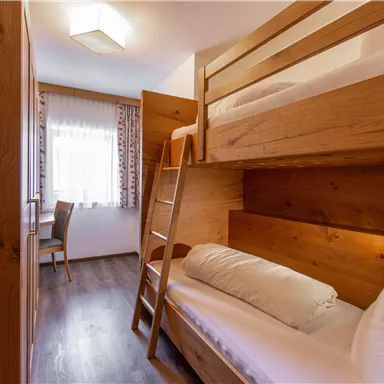 A cozy room with a wooden bunk bed and a single bed. A table and a chair complete the furnishings, and the window lets in a lot of light.