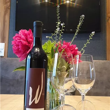 Eine Flasche Wein steht auf einem Tisch, umgeben von zwei Weingläsern und bunten Blumen. Im Hintergrund ist ein Fernseher zu sehen.