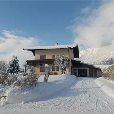 Ein gemütliches Haus im Schnee mit einer schneebedeckten Auffahrt. Im Hintergrund sind verschneite Berge und ein klarer Himmel zu sehen.