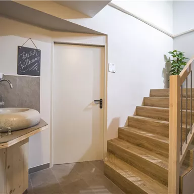 Ein moderner Eingangsbereich mit einer Holztreppe und einem eleganten Waschbecken. An der Wand hängt eine kleine Tafel und es gibt einige Pflanzen zur Dekoration.